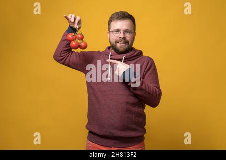 Un homme à barbe souriant et satisfait avec des verres tient une branche de tomates mûres rouges à la main et pointe vers elle avec l'index. Banque D'Images