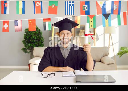 Bonne université internationale en ligne diplômé assis à la table, titulaire d'un diplôme et souriant Banque D'Images