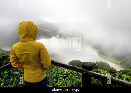 Garçon en blouson de pluie jaune debout parmi les plantes, regardant au lac cratère Lagoa Funda das Lajes dans le brouillard, île de Flores, Açores, Portugal Banque D'Images