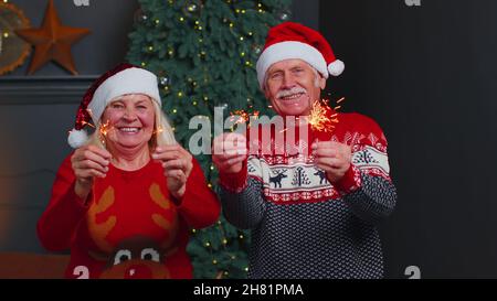 Couple de familles âgées en chandails festifs tenant des lumières bengale appréciant la veille de Noël dans la chambre à la maison Banque D'Images