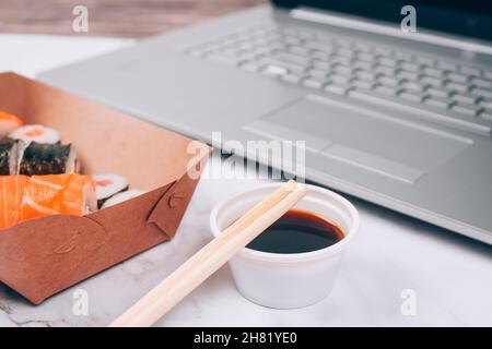 Différents rouleaux de sushis avec saumon dans une boîte de papier de livraison sur fond de table en marbre blanc avec ordinateur portable - futomaki, uramaki et hosomaki. Commande en ligne Banque D'Images