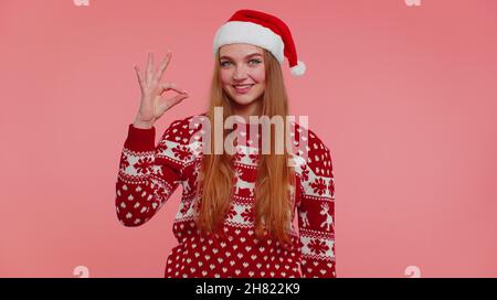 Fille adulte dans le chandail de Noël regardant approvement à l'appareil photo montrant le geste ok, comme signe positif Banque D'Images
