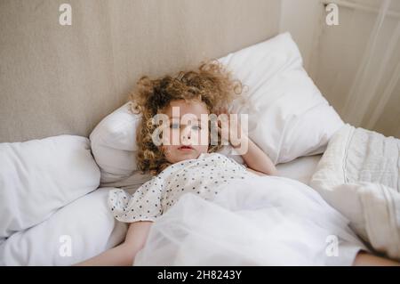 Jolie petite fille aux cheveux bouclés couchée dans un lit sur un oreiller blanc.Le concept de l'enfance. Banque D'Images