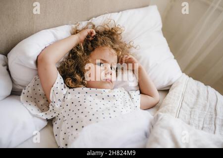 Jolie petite fille aux cheveux bouclés couchée dans un lit sur un oreiller blanc.Le concept de l'enfance. Banque D'Images