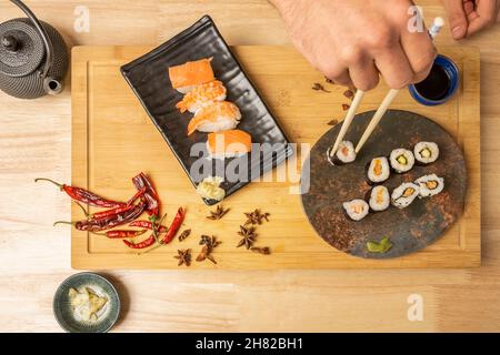 Main d'un jeune homme tenant un rouleau maki d'une assiette à côté d'autres plats de sushi avec nigiri de saumon et piments rouges chauds Banque D'Images