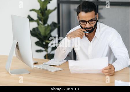 Homme d'affaires indien barbu, sérieux et concentré, dans une tenue officielle, assis à une table au bureau, étudiant des documents, analysant un rapport et des tableaux financiers Banque D'Images