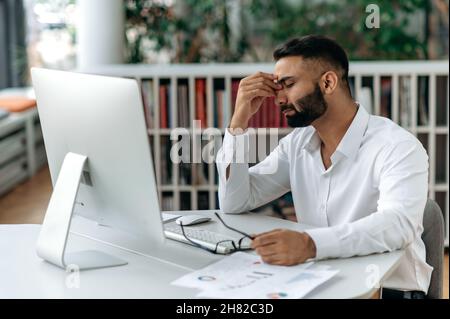 Un Indien fatigué épuisé, propriétaire de l'entreprise ou employé de bureau, s'assoit au bureau avec les yeux fermés, se frotte les yeux, se sentant mal de tête, stress et fatigue du travail supplémentaire, besoin de repos et de pause Banque D'Images