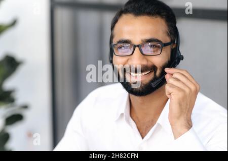 Gros plan d'un homme d'affaires indien intelligent, d'un opérateur de centre d'appels ou d'un consultant, portant des lunettes, utilisant un casque pour les conférences téléphoniques, regardant l'appareil photo et souriant amical Banque D'Images