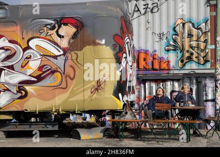 Deux hommes ayant un verre à côté du graffiti pulvérisé vieille voiture de train dans la zone de projet de culture de Bahnwaerter Thiel, Munich, Bavière, Allemagne. Banque D'Images