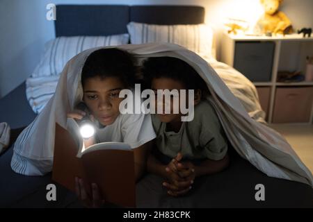 Curieux Afro-américains enfants couchés ensemble sur le lit et livre de lecture avec lampe de poche la nuit Banque D'Images