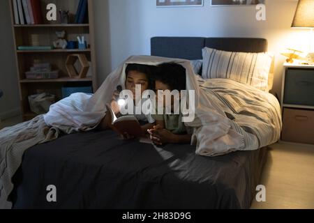 Soeur afro-américaine tenant une lampe de poche et un livre de lecture à frère pendant qu'ils sont au lit la nuit Banque D'Images