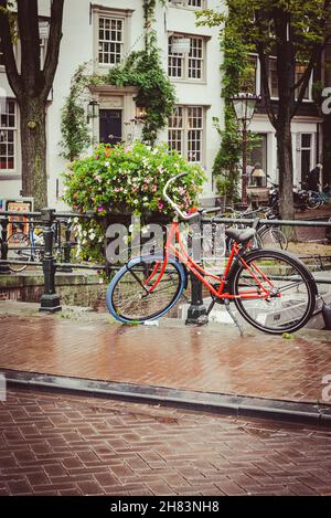 Un vélo rouge se trouve à l'extérieur, stationné sur un pont au-dessus d'un canal dans la ville d'Amsterdam, tandis qu'il y a une pluie torride qui fait mouiller les rues pavées Banque D'Images