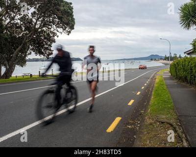 AUCKLAND, NOUVELLE-ZÉLANDE - 07 juillet 2021 : deux personnes qui font du jogging et du vélo sur une route longue exposition lors d'une journée de pluie à Auckland, Nouvelle-Zélande Banque D'Images