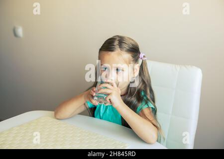 Belle petite fille boit de l'eau propre d'un verre transparent. Banque D'Images