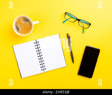 Vue de dessus d'un bureau jaune avec un ordinateur portable vide, un stylo, un téléphone, des lunettes et du café Banque D'Images