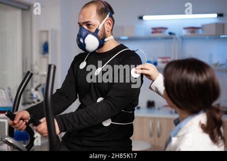 Médecin spécialiste médecin mettant des électrodes capteur sur le sportif avec masque de surveillance des données de l'EGK dans le laboratoire d'examen médical.Athlète homme en train de courir sur un entraîneur sans finale travaillant à l'endurance du corps Banque D'Images