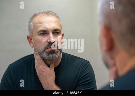 Homme d'âge moyen regardant dans le miroir de la salle de bains touchant sa barbe en pensant à la couper Banque D'Images