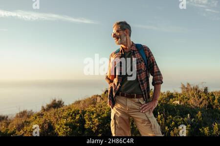 Randonneur insouciant qui regarde gaiement sur une colline.Joyeux randonneur mûr souriant en se tenant au sommet d'une colline avec un sac à dos.Le dos d'un homme aventureux Banque D'Images