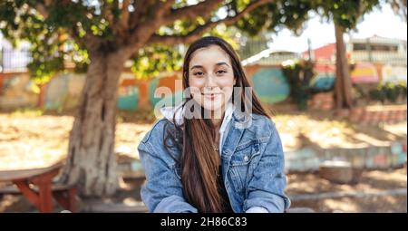 Adolescente confiante regardant l'appareil photo avec un sourire.Gaie brune fille portant une veste en denim à l'extérieur de la ville.Sittin pour jeunes femmes Banque D'Images