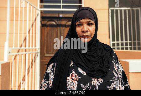 Femme musulmane confiante regardant la caméra tout en se tenant devant sa maison pendant la journée.Une femme d'âge moyen confiante porte un hijab et Banque D'Images