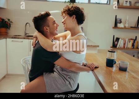 Couple étant confortable dans la cuisine.Une jeune femme affectueuse embrasse son petit ami tout en étant assise sur ses genoux.Couple romantique de jeunes reines souriant à ea Banque D'Images