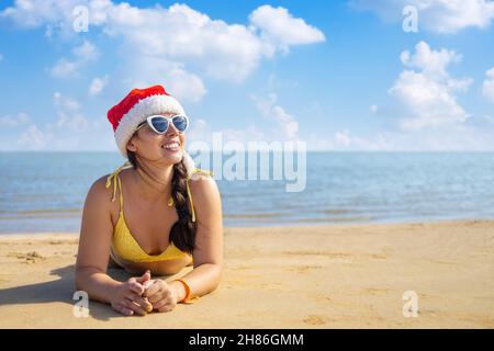 femme souriante en chapeau de père noël et lunettes de soleil couchés sur une plage de sable Banque D'Images