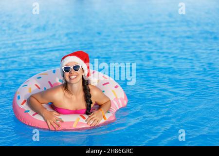 portrait de la jeune femme heureuse en chapeau de père noël et lunettes de soleil se détendant sur l'anneau gonflable dans la piscine Banque D'Images