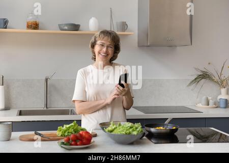 Bonne femme mature propriétaire d'une maison utilisant un téléphone portable dans la cuisine Banque D'Images
