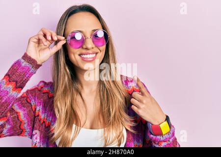 Belle femme hispanique portant le style bohème et hippie avec des lunettes de soleil roses souriant heureux pointant avec la main et le doigt Banque D'Images