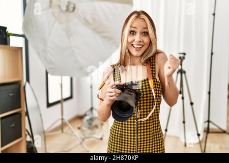 Jeune photographe caucasien tenant un appareil photo professionnel dans un studio de photographie criant fier, célébrant la victoire et le succès très excité avec Banque D'Images