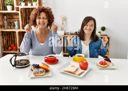 Famille de mère et de fille de syndrome de Down assis à la maison manger petit déjeuner excité pour le succès avec les bras levés et les yeux fermés célébrant la victoire Banque D'Images