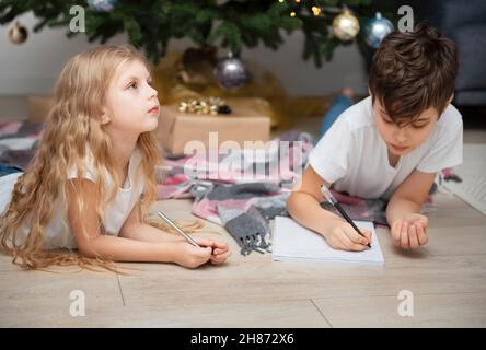 Enfants - un garçon et une fille écrivent des lettres au Père Noël près de l'arbre de Noël dans le salon Banque D'Images