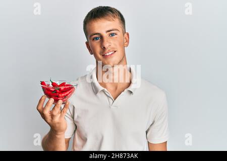 Jeune homme caucasien tenant un bol de poivre rouge en regardant positif et heureux debout et souriant avec un sourire confiant montrant des dents Banque D'Images