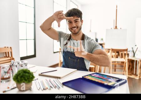 Jeune homme hispanique dans un studio d'art souriant faisant cadre avec les mains et les doigts avec le visage heureux. Créativité et concept de photographie. Banque D'Images