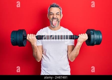 Beau homme d'âge moyen avec des cheveux gris portant des vêtements de sport en utilisant des haltères collant la langue dehors heureux avec l'expression drôle. Banque D'Images