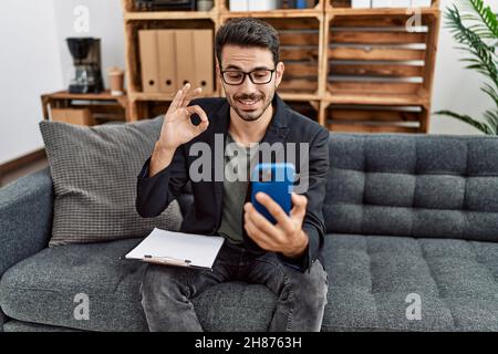 Jeune psychologue hispanique homme faisant la thérapie sur appel vidéo avec smartphone faisant signe ok avec les doigts, souriant sympathique gestante excellent symbole Banque D'Images