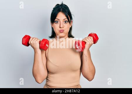 Jeune femme hispanique portant des vêtements de sport utilisant des haltères puffing joues avec visage drôle. Bouche gonflée avec de l'air, attrapant l'air. Banque D'Images