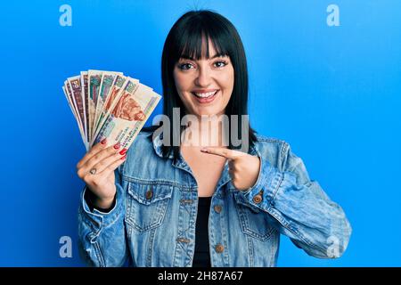 Jeune femme hispanique portant des billets en livres égyptiennes souriant, souriante, pointant du doigt et de la main Banque D'Images