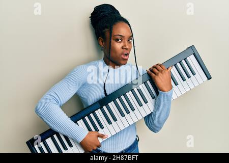 Femme afro-américaine avec cheveux tressés tenant le clavier de piano dans le visage de choc, regardant sceptique et sarcastique, surpris par la bouche ouverte Banque D'Images