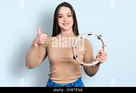 Jeune fille hispanique jouant tambourine souriant heureux et positif, pouce vers le haut faisant excellent et signe d'approbation Banque D'Images