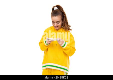 Jolie belle souriante gaie mignonne avec des cheveux foncés avec une queue de cheval dans un élégant jaune tracksuit avec des rayures vertes joue un téléphone mobile avec Banque D'Images