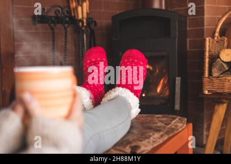 La femme boit du café chaud devant la cheminée et réchauffe ses pieds avec des chaussettes en laine.Détente dans le salon.Plaisir de la vie domestique Banque D'Images