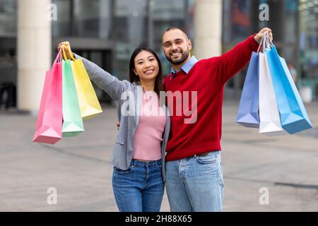 Un couple millénaire et diversifié se tenant près du centre commercial, tenant des sacs de shopper dans des mains tendues, souriant à l'appareil photo Banque D'Images