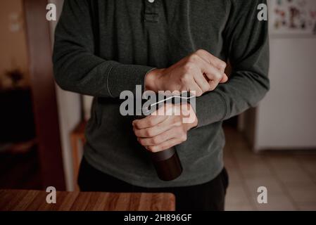 Jeune hippster millénaires de course mixte homme meulant le café à l'aide d'un petit moulin à burr manuel dans la cuisine à la maison avec éclairage moody. Banque D'Images