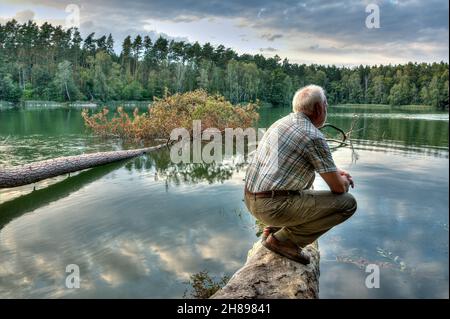 Dans la dernière lumière du soleil couchant, un vieil homme s'assoit sur une bûche sur un lac solitaire et jouit du silence et du calme de l'heure du soir dans la nature. Banque D'Images