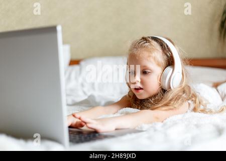 Blanche blonde enfant fille utiliser un ordinateur portable dans le casque pour faire des devoirs regarder des vidéos ou des dessins animés.Fille assise sur des études de lit par appel vidéo Banque D'Images