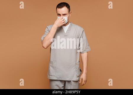 Un jeune thérapeute dans un masque jetable sur fond beige.Jeune médecin virologue portant un masque de protection et un uniforme médical isolé sur Banque D'Images