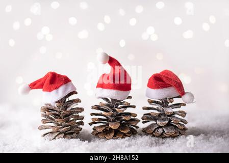 Trois cônes de pin dans un chapeau de père Noël rouge comme un arbre de Noël avec des lumières de bokeh dans la neige sur un fond blanc, concept de Noël. Banque D'Images