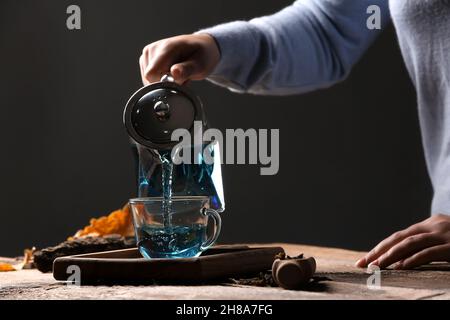 Femme versant le thé de fleur de pois de papillon de la théière dans une tasse sur la table sur fond sombre Banque D'Images