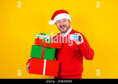Portrait d'un homme d'âge latin tenant une carte de crédit pour faire du shopping à Noël portant un chapeau de père noël sur fond jaune au Mexique en Amérique latine Banque D'Images
